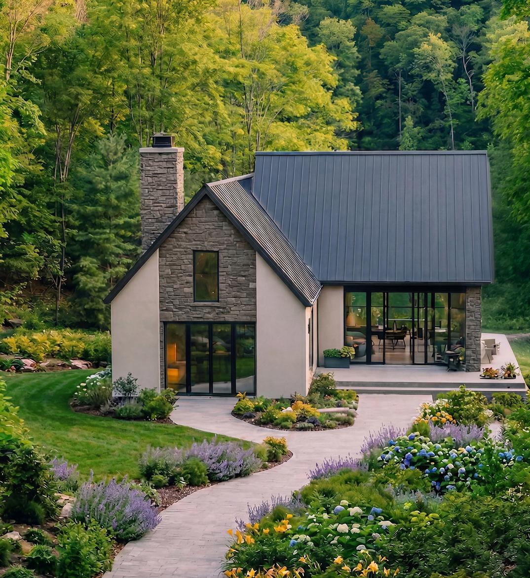 Aerial view of a modern farmhouse-style home with stucco and stone siding, a dark metal roof, and a stone chimney, surrounded by colorful gardens with lavender, hydrangeas, and daylilies. A curved paver path leads to the entrance. Dense green forest in the background.