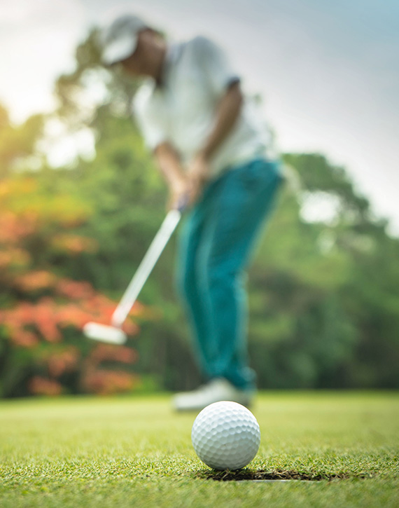 property-slider-Golf Course Lots A close-up of a golf ball on a green fairway with a golfer lining up a putt in the blurred background, surrounded by colorful autumn trees.