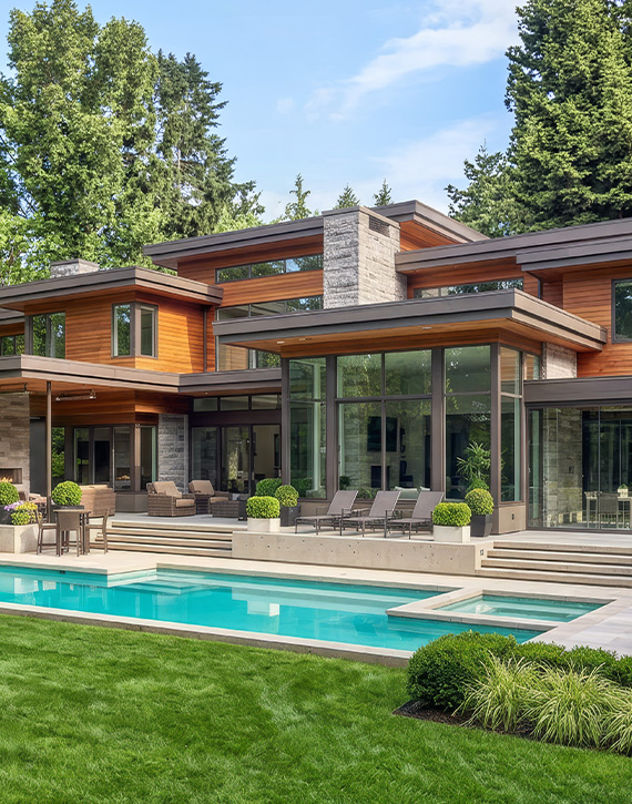 property-slider-Interior Waterfront Lots Exterior of a two-story contemporary home featuring warm wood siding, natural stone columns, large black-framed windows, and lush landscaping with a paved walkway. Tall evergreen trees in the background.